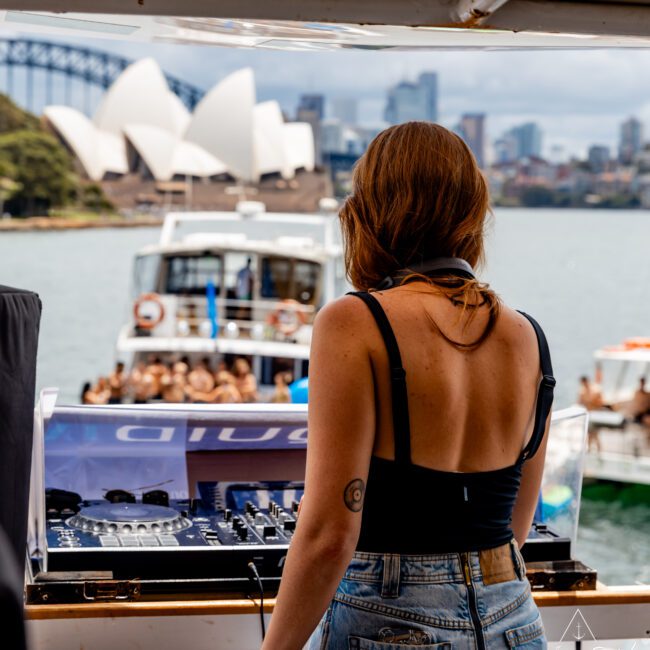 A DJ with long hair, wearing a black top and jeans, performs on a boat with music equipment. A crowd on another boat listens in the background. The Sydney Opera House and Harbour Bridge are visible across the water.