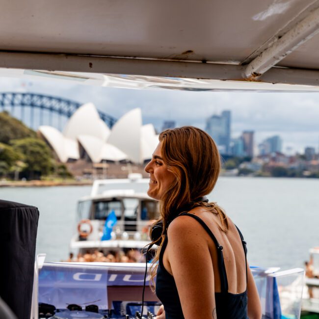 A woman DJ stands on a boat deck, wearing headphones and a black top. The Sydney Opera House and Harbour Bridge are visible in the background. The scene is vibrant, with blue skies and water, creating a lively atmosphere.