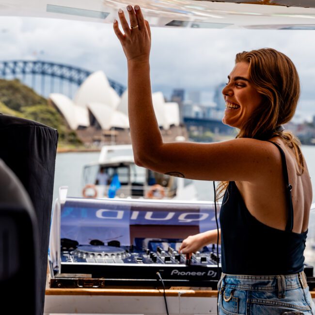 Woman DJing on a boat, smiling and interacting with the crowd. She is wearing a black top and jeans, with DJ equipment in front of her. In the background, the Sydney Opera House and Harbour Bridge are visible under a cloudy sky.