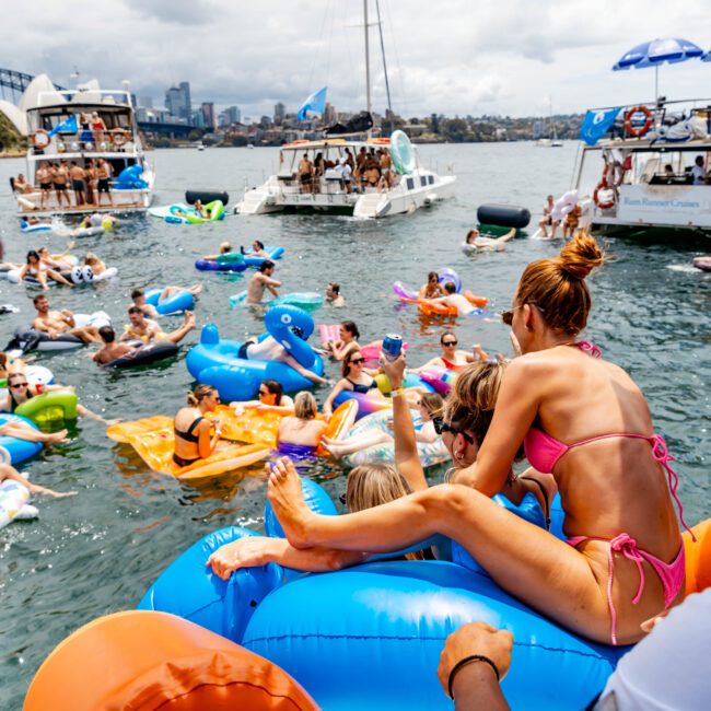 People enjoying a summer party on a sunny day, floating on inflatables in a body of water. Boats are anchored nearby, with people sitting and standing on them. A woman in a pink bikini sits on an inflatable, holding a child. City skyline in the distance.