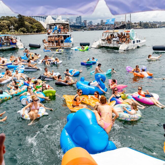 A lively scene of people enjoying a sunny day on colorful inflatable toys in a body of water. Boats are anchored nearby, and the background features a city skyline with a bridge. A person films the event while others relax and socialize in the water.