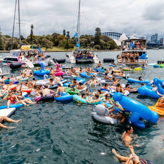 A large group of people relaxing on colorful inflatables and floating platforms in the water near several yachts. The background features trees, city buildings, and a bridge under a cloudy sky.