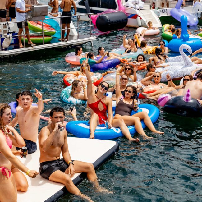 A lively scene of people enjoying a party on the water. They are lounging on inflatables and a floating platform, with drinks in hand. There are various colorful floaties, a nearby boat, and people standing and sitting around, all under a sunny sky.