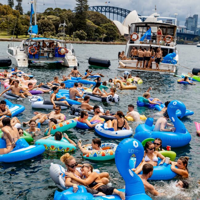 A lively scene on the water with people on inflatable floats, surrounded by yachts. The Sydney Opera House and Harbour Bridge are visible in the background. Participants are socializing and enjoying the sunny day.