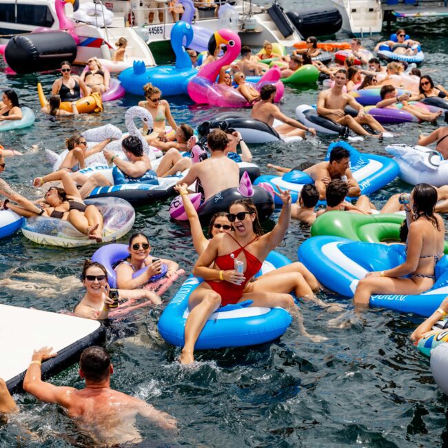 A group of people enjoying a sunny day on colorful inflatable floats in the water. They are surrounded by yachts and waving, holding drinks, and smiling. There are flamingo and unicorn inflatables, creating a festive, lively atmosphere.