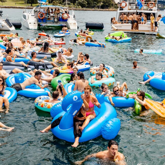 A lively water scene features numerous people in swimsuits on colorful inflatable floats in a bay. Boats are in the background, with a bridge and iconic building visible under a clear sky.