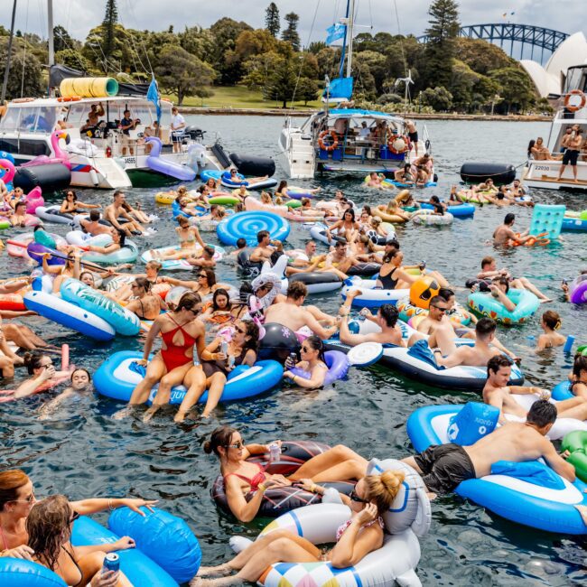 A vibrant gathering of people relaxing on colorful inflatable floats in the water. Boats are nearby, and the background features trees and part of a bridge. The scene exudes a lively and festive atmosphere.