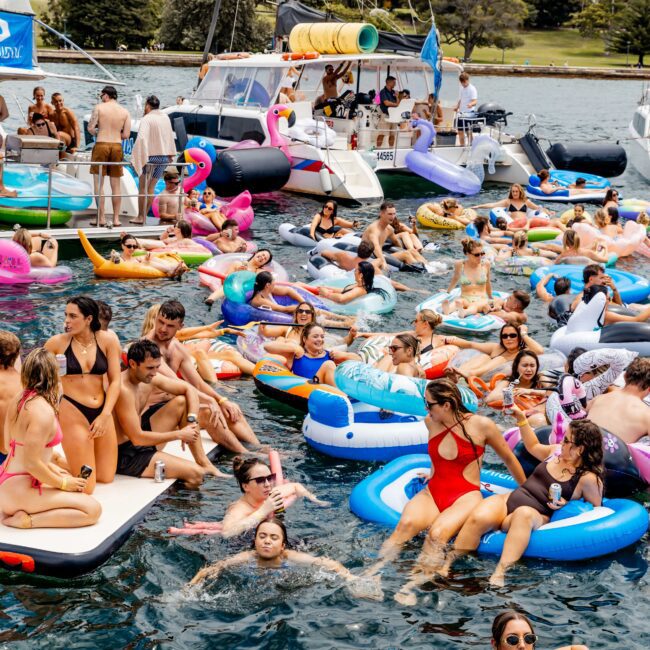 A lively scene of people enjoying a party on a lake. They are floating on colorful inflatables and a catamaran is docked nearby. Many are wearing swimwear, and the atmosphere is festive with laughter and socializing under a partly cloudy sky.