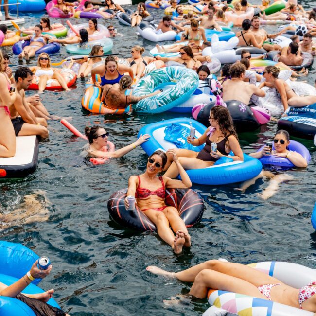 A lively gathering of people on inflatable rafts and tubes in a body of water, surrounded by boats. They're enjoying a sunny day, with food, drinks, and various colorful floaties, including a pizza and unicorn shape.