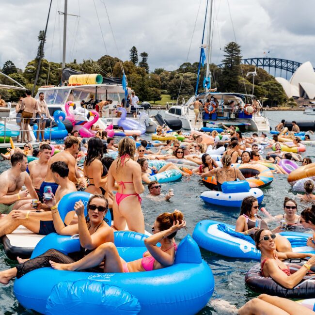 A lively gathering of people on inflatable floats in the water, surrounded by boats. Participants in swimsuits enjoy sunny weather, with some holding drinks. A bridge can be seen in the background.