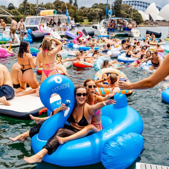 People enjoying a sunny day on colorful inflatable rafts and boats in a bustling harbor. A large bridge and iconic white sails of the Sydney Opera House are visible in the background. The scene is lively and festive.