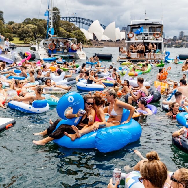 A lively scene with many people floating on inflatable tubes in a body of water. A cruise boat is in the background, with Sydney Opera House and Harbour Bridge visible. The sky is cloudy, and participants are enjoying the party atmosphere.