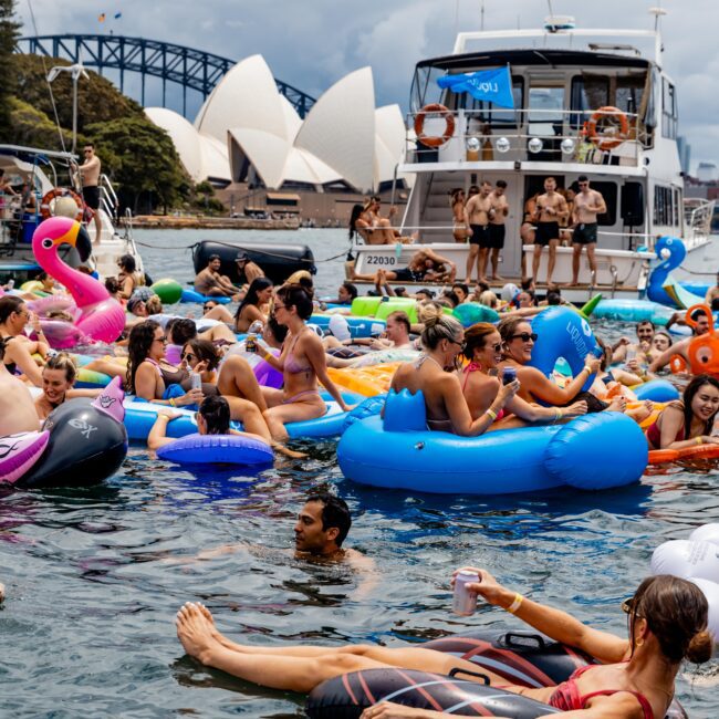 A lively scene of people enjoying a boat party in a harbor. Many float on colorful inflatables in the water. The iconic Sydney Opera House and Harbour Bridge are visible in the background under a cloudy sky.