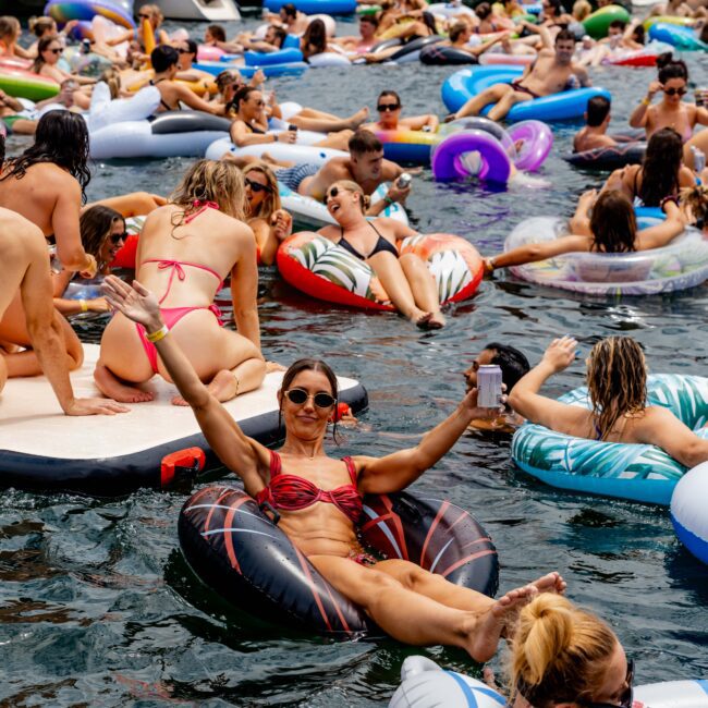 A crowded scene on the water with people relaxing and socializing on inflatable rafts and floats. Many are wearing swimsuits and sunglasses, enjoying a sunny day. Boats are anchored nearby in the background.