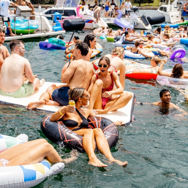 People enjoying a sunny day on a lake, lounging on inflatables and floating platforms. Boats are anchored nearby, and the mood is festive. The scene is lively with people in swimsuits relaxing and talking.