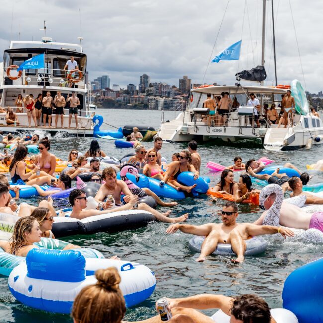 A crowded gathering of people enjoying a sunny day on the water, lounging on colorful inflatable floats. Boats with more people are anchored nearby. The city skyline is visible in the background under a partly cloudy sky.