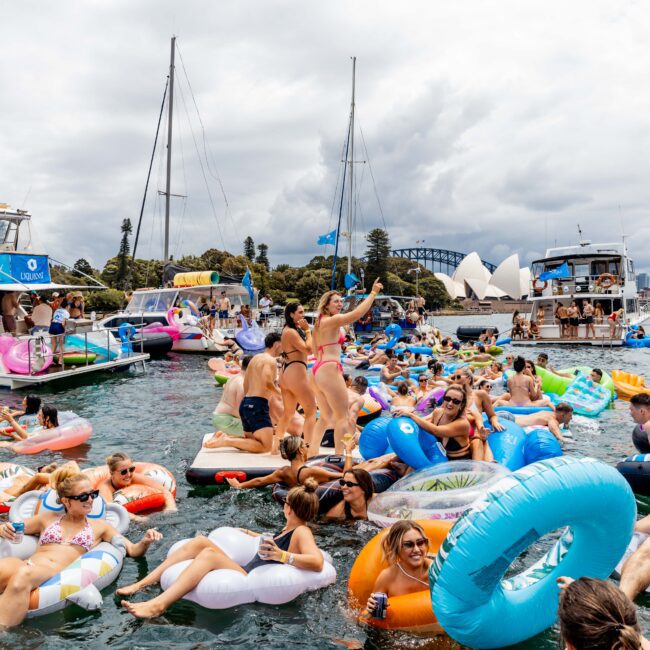 A large group of people enjoying a sunny day on inflatable rafts in the water, surrounded by boats. The iconic Sydney Opera House and Sydney Harbour Bridge are visible in the background, under a cloudy sky.