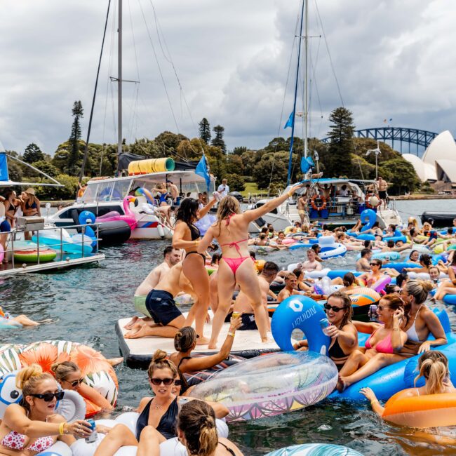 A large group of people enjoy a party on the water, floating on colorful inflatable rafts and tubes. Sailboats and the iconic Sydney Opera House and Harbour Bridge are visible in the background. The sky is cloudy.