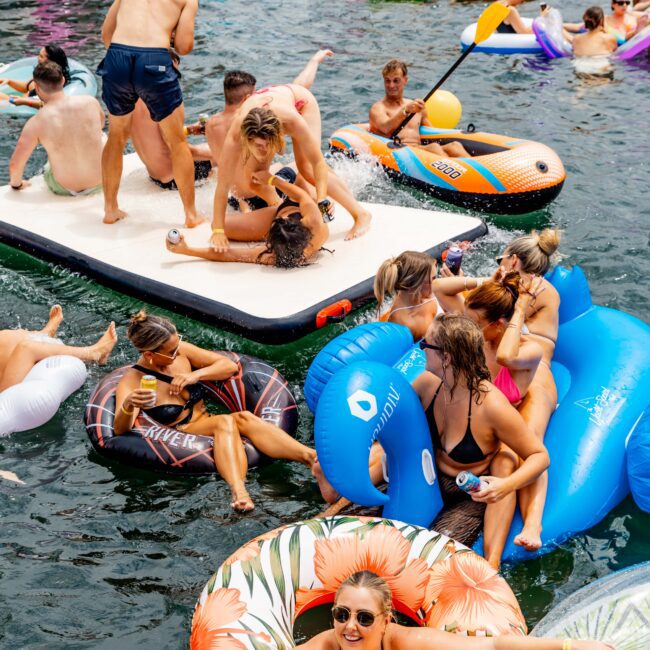 A lively scene of people enjoying a party on inflatable floats in the water. Some are lounging, while others are engaging in playful activities. Colorful floats and a floating platform are visible, with boats in the background.