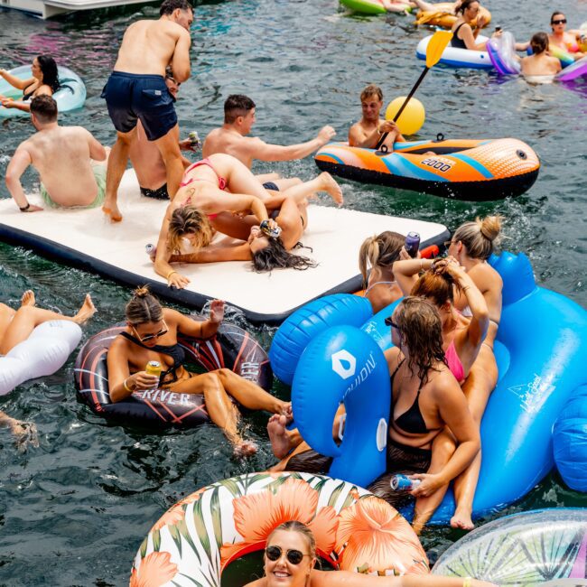 A lively scene at a lake party with people lounging on colorful inflatables and paddleboards. Some are in the water, while others relax on a floating platform. Brightly colored tubes and boats are scattered around, creating a fun, festive atmosphere.