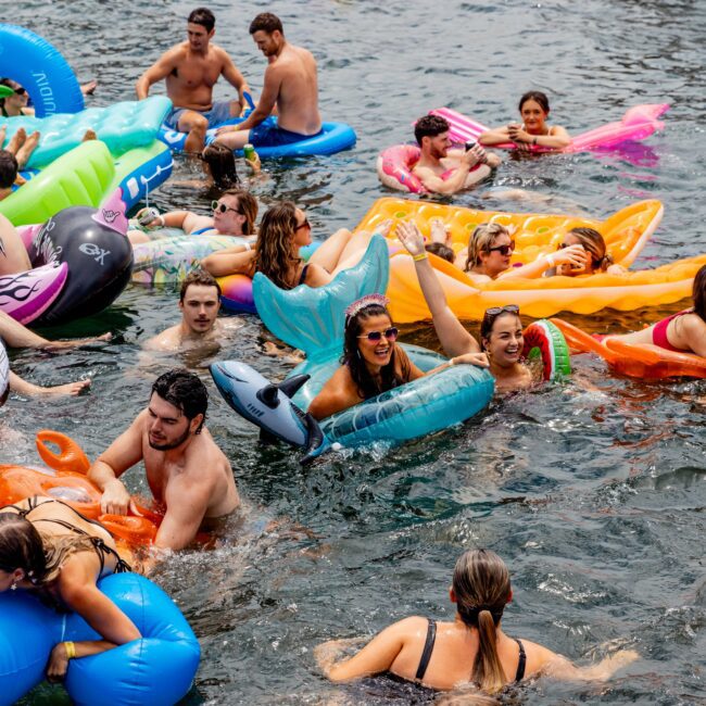 A group of people enjoying a water party, floating on colorful inflatable rafts and tubes in a lake or ocean. There's a boat in the background and everyone is smiling and having fun in the sun.