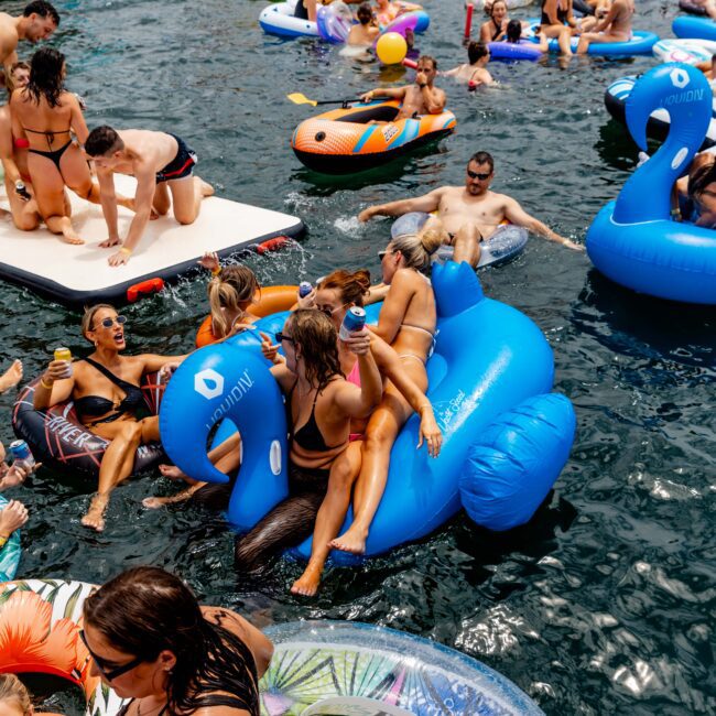 People enjoying a sunny day on a lake with inflatable floaties, including a large blue swan and colorful rings. Boats are in the background, and the water is filled with swimmers and people lounging on the floats.