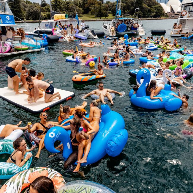 A lively party scene on the water with people enjoying various colorful inflatable floats. Surrounding yachts provide a backdrop, while many partygoers relax and chat in a festive atmosphere under a clear sky.