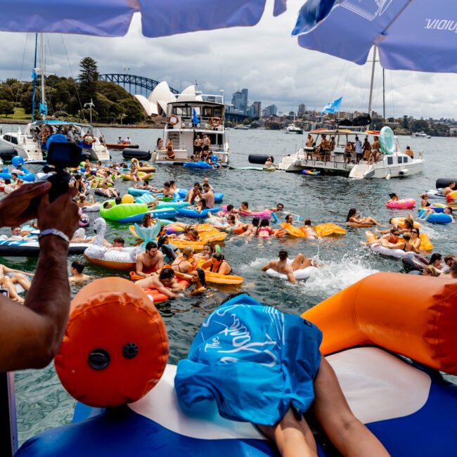 A lively scene of people on colorful inflatable pool floats enjoying a party in the water. Umbrellas offer shade, and boats are docked nearby. The sky is cloudy, with a distant view of a city skyline and a bridge.