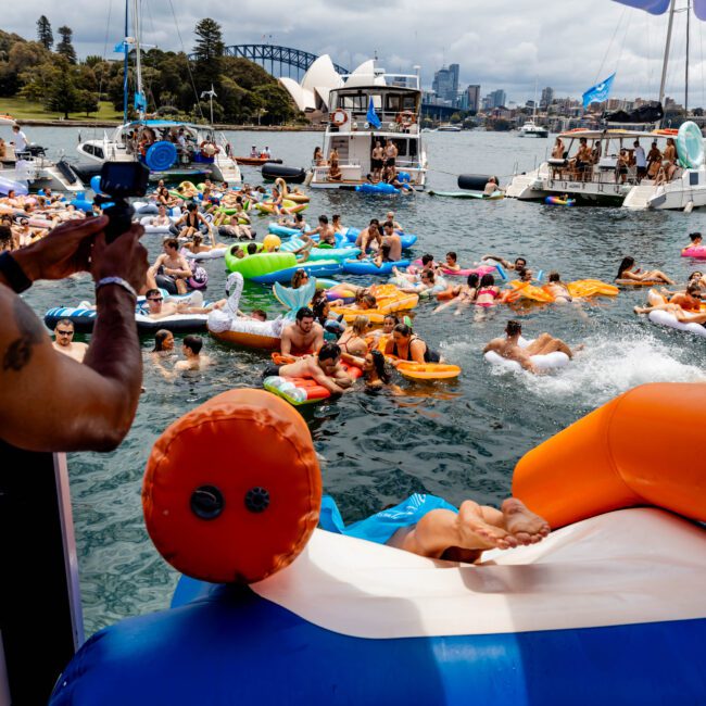 A lively scene of people enjoying a summer day on the water, surrounded by inflatable floats and boats. Others socialize and relax under umbrellas. The backdrop features a cityscape and a bridge, indicating a festive waterfront gathering.