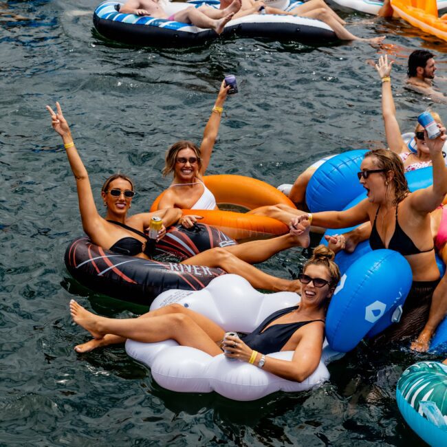 A crowded group of people enjoying a day on the water, floating on various inflatable rings and loungers. They are laughing, raising their arms, and holding drinks, creating a lively and festive atmosphere.