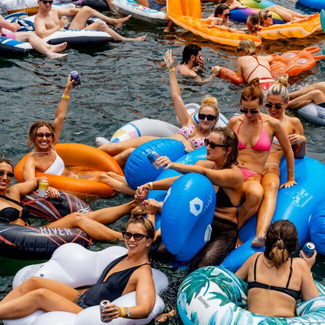 A group of people in swimsuits are relaxing and enjoying drinks on colorful inflatable floaties in a body of water. They are smiling and making peace signs. The scene is lively and joyful, with sunlight reflecting on the water.