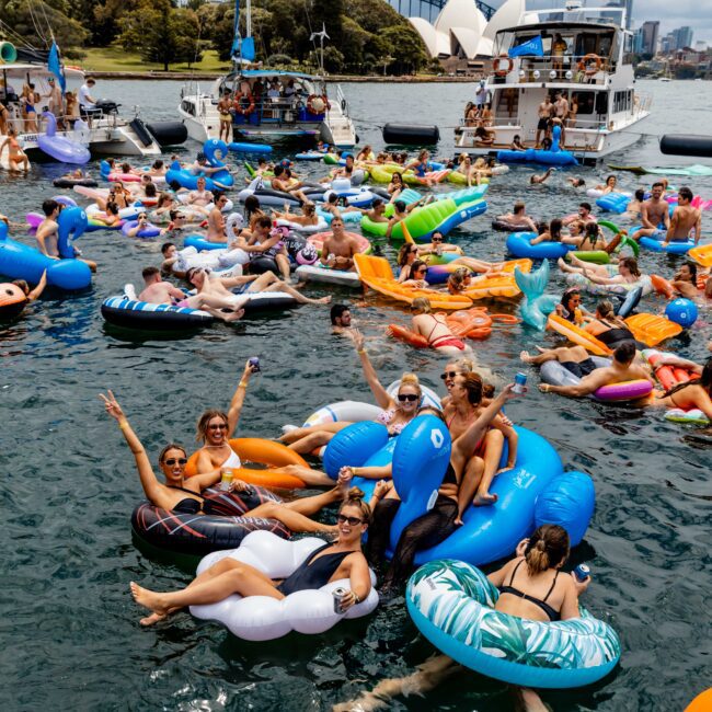 A lively scene of people floating on inflatable pool toys in a harbor. Various colorful floats, including unicorns and swans, fill the water. Boats are nearby, with distant views of a city skyline and an iconic white structure.