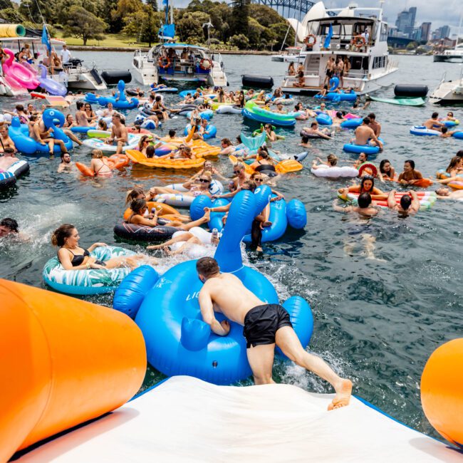 A lively scene of people enjoying a day on the water with numerous inflatables of various shapes and colors. They are surrounded by boats, and a cityscape with a partially visible bridge is in the background under a cloudy sky.