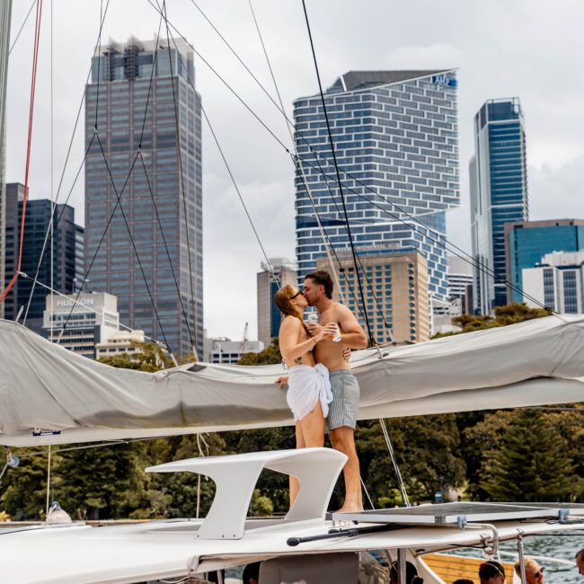 A couple embraces on a yacht under a blue flag, with a city skyline in the background. Below, a group of people is enjoying the boat party, some chatting and wearing casual summer clothes. Skyscrapers and trees are visible.