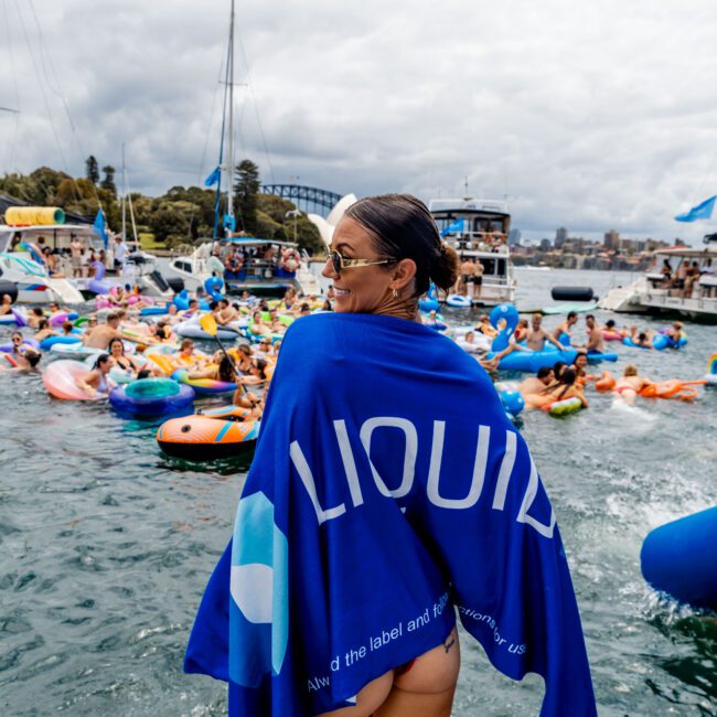 A person with sunglasses stands on a dock draped in a blue towel with a "LIQUID" logo. In the background, numerous people in colorful inflatable rafts are gathered on the water under a partly cloudy sky.