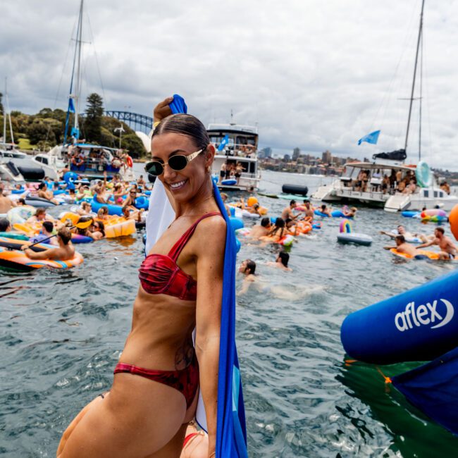 A woman in a red bikini poses on a boat's edge, holding a blue and white flag. She is surrounded by a lively crowd on inflatables in the water, with boats and a cloudy sky in the background.