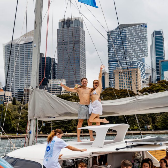 A group of people enjoying a yacht outing near a city skyline with modern skyscrapers. Two people stand on a raised platform, smiling and raising their hands. Others are relaxed on the deck. A blue flag flutters above.