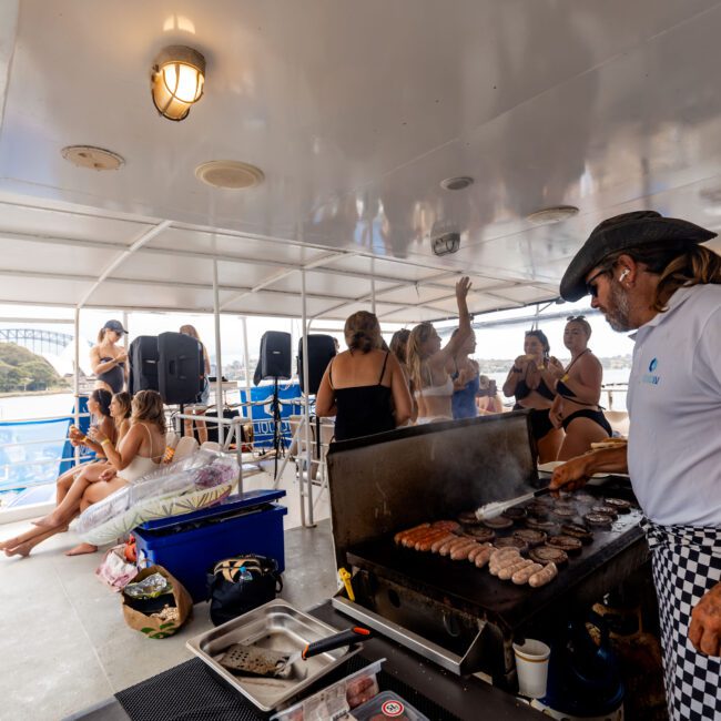 A man grills burgers on a boat while several people in swimwear are in the background. Some are sitting, chatting, and enjoying the view of the water. A bridge is visible in the distance. The atmosphere is lively and relaxed.