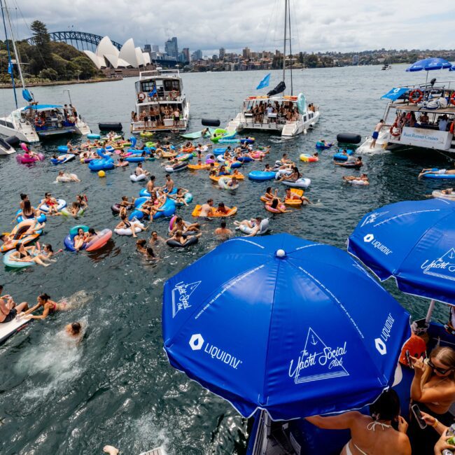 A vibrant boat party on a sunny day in a harbor. People are on colorful inflatable floats and boats nearby. Large blue umbrellas in the foreground and the iconic Sydney Opera House and skyline are visible in the background.