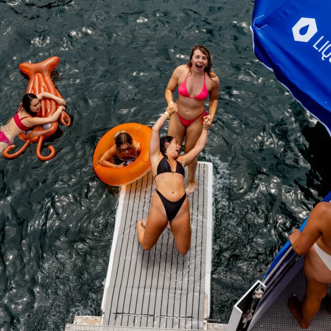 Two women in swimsuits are joyfully dancing on a metal platform extending over water, with others swimming nearby. An inflatable pool toy resembling an octopus is floating, and a blue beach umbrella is partially visible.