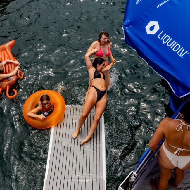 People are enjoying a day at the lake. A woman in a black bikini steps onto a floating dock, while others swim nearby. An orange float and a blue umbrella with "Liquid IV" text are visible.