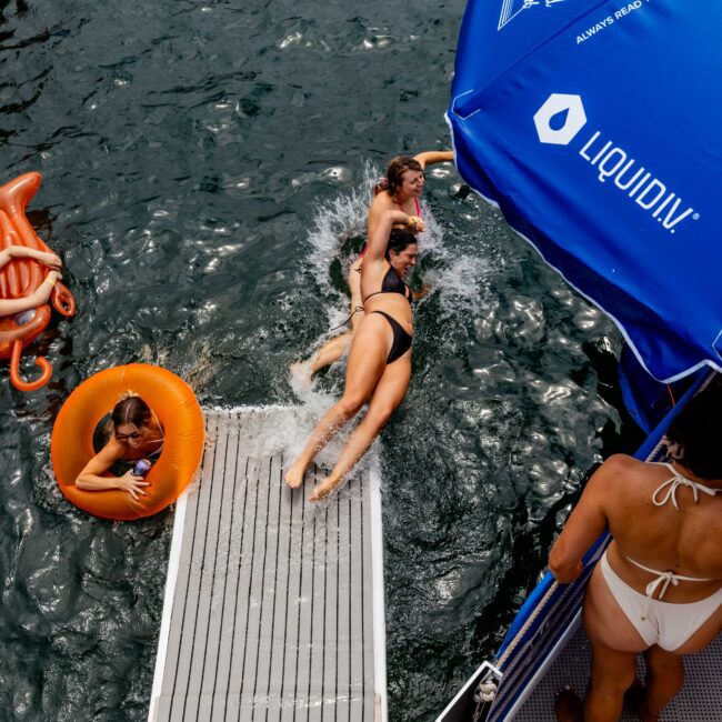 Three women in swimsuits descend a ramp into the water near a dock, with one using an orange float. Others swim nearby. A blue umbrella is visible on the right. The scene is lively, with people enjoying a day by the water.