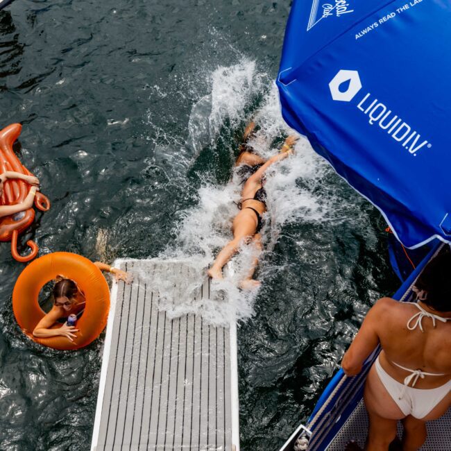 A person slides into the water from a boat using a wide slide, making a splash. Another person in the water holds an orange float ring. The boat has a blue canopy with white logos. Other swimmers are visible in the background.