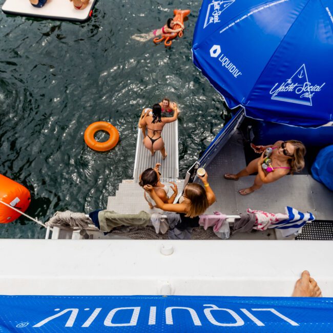 Overhead view of people enjoying a party on a yacht. The scene includes people swimming, lounging on floats in the water, and relaxing on the steps of the boat. There are branded blue umbrellas and a large branded banner visible.