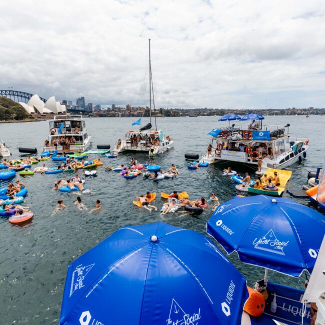 People enjoying a boat party on a harbour, with numerous colorful inflatables in the water. The Sydney Opera House and Harbour Bridge are visible in the background. Blue umbrellas labeled "Lite & Social" are in the foreground.