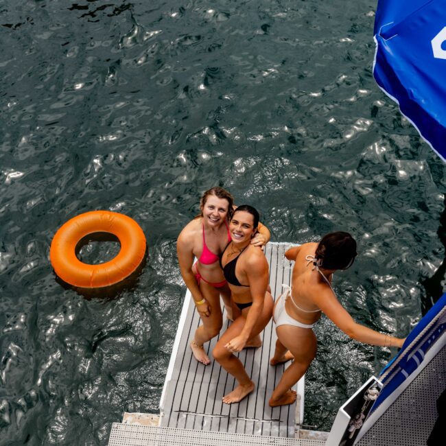 Three people in swimsuits stand on a boat's edge, smiling at the camera. They are next to a large floatation device in the water. The scene is lively, with others also enjoying the water nearby.