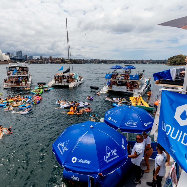 A lively party scene on a body of water with people on colorful inflatables and boats. Blue umbrellas with "Liquid I.V." branding are visible. The Sydney Opera House and Harbour Bridge are in the background under a partly cloudy sky.