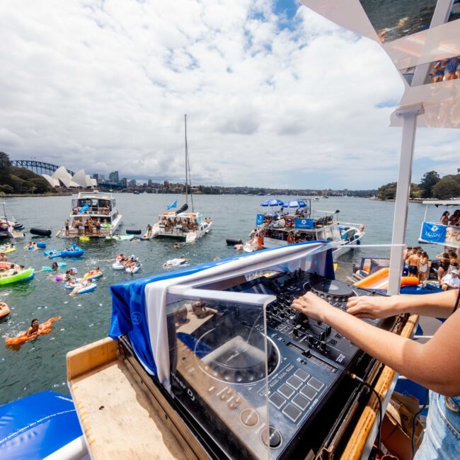 A DJ plays music on a boat overlooking a lively scene of people on colorful inflatables in the water. Nearby boats are anchored, and the Sydney Opera House and Harbour Bridge are visible in the background under a cloudy sky.