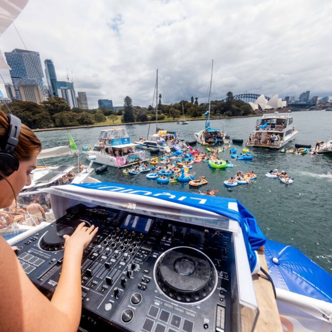 A DJ is performing on a boat deck, overlooking a lively scene of people on inflatables and in the water. Several other boats are in the vicinity. The city skyline and famous architectural landmarks are visible in the background.