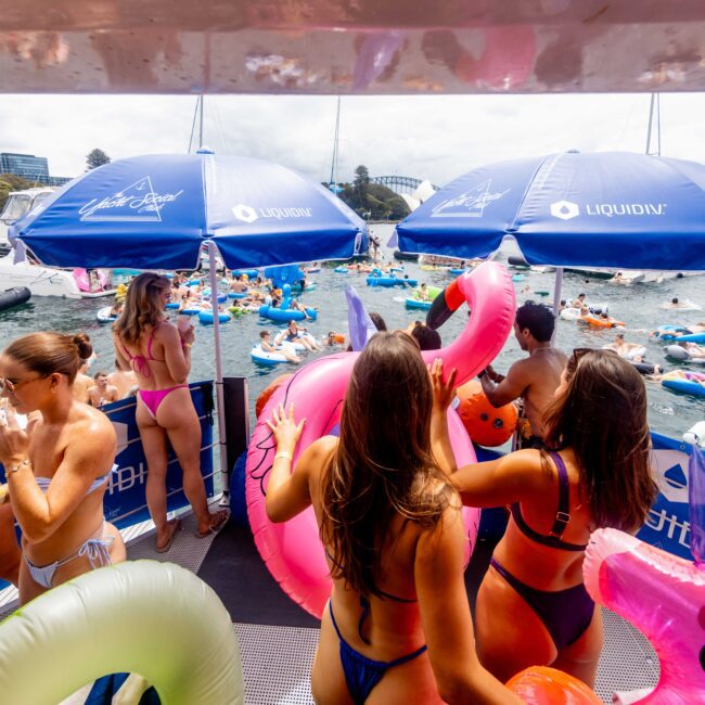 People in swimsuits enjoying a lively boat party on a sunny day. The scene features water floats and inflatable pool toys. Blue Liquid DJ umbrellas provide shade. The background includes boats and a waterfront skyline.